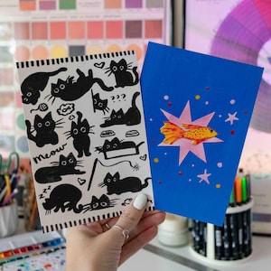 May include: Two greeting cards. One card features black cat illustrations with the word "meow". The other card is blue with a gold fish illustration. The background includes color charts and art supplies.