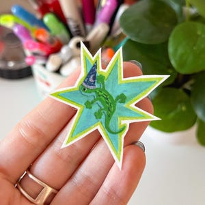 May include: A hand-drawn sticker featuring a green lizard wearing a wizard's hat, set against a blue starburst design with a green border. The lizard has a textured appearance, and the hat is blue with white stars.