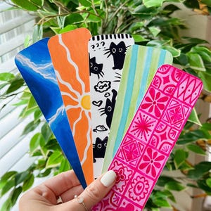 May include: A hand holds a fan of five colorful bookmarks. The bookmarks feature various designs, including a blue watercolor sky, an orange sun, black cat silhouettes, green and blue stripes, and a pink floral pattern. The background is a green plant.