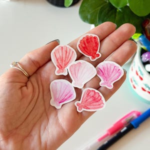 Puede incluir: Una mano sostiene siete pegatinas de conchas marinas. Las pegatinas están pintadas con acuarela en tonos rosa y rojo. Presentan patrones detallados de conchas y varían en tamaño y forma. El fondo incluye bolígrafos, una planta y útiles de arte.