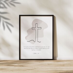 Könnte beinhalten: Ein druckbares Wandbild mit einer minimalistischen Linienzeichnung eines Kreuzes vor einem zartrosa Aquarellhintergrund. Der Text "Denn so sehr hat Gott die Welt geliebt, dass er seinen einzigen Sohn hingab, damit jeder, der an ihn glaubt, nicht verloren geht, sondern ewiges Leben hat. - Johannes 3,16 -" ist unter dem Kreuz gedruckt.