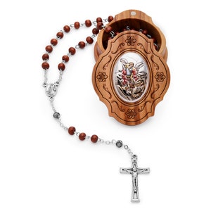 Puede incluir: Un rosario de madera marrón con una cruz de plata y una medalla de plata con la imagen de un ángel. El rosario está en una caja de madera marrón con un diseño tallado.