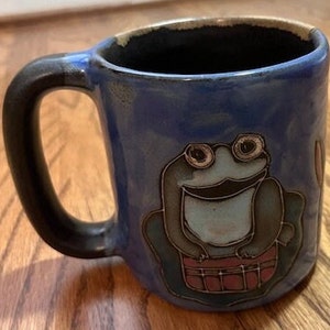 May include: Blue ceramic mug with a frog design. The frog is sitting on a red and white striped barrel. The mug has a black handle.