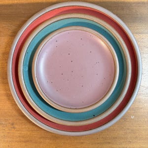 May include: A stack of three ceramic plates in varying sizes. The largest plate has a red rim, the middle plate has a teal rim, and the smallest plate is pink. All plates have a speckled texture.