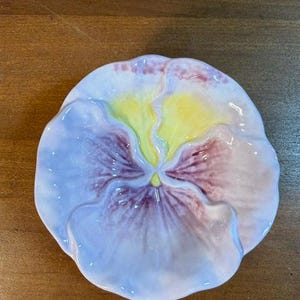 May include: A ceramic dish shaped like a pansy flower with wavy, scalloped edges. The petals feature a gradient of soft lavender and light blue, transitioning into a deep purple center with a bright yellow heart at the very middle of the bloom.