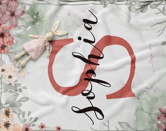 May include: A white baby blanket with a floral design in shades of pink, green, and gray. The blanket features a large red letter "S" and the name "Sophia" written in black cursive script.