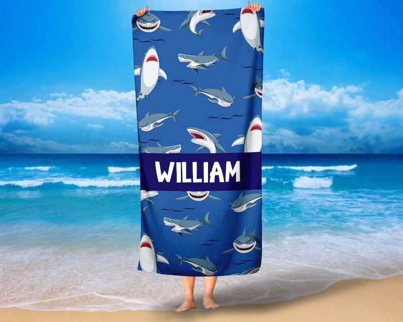 May include: A blue beach towel with a white shark pattern and the name "WILLIAM" printed in white on a navy blue stripe.