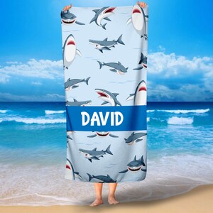 May include: A blue beach towel with a white background and a pattern of cartoon sharks. The towel has a blue stripe across the middle with the name "DAVID" printed in white.