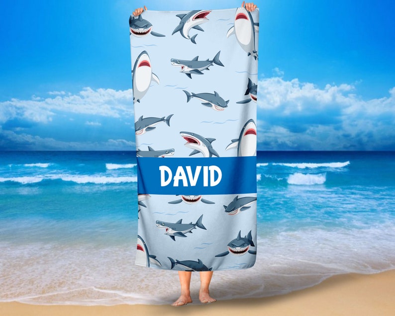 May include: A blue beach towel with a white background and a pattern of cartoon sharks. The towel has a blue stripe across the middle with the name "DAVID" printed in white.