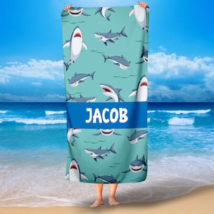 May include: A turquoise beach towel with a repeating pattern of cartoon sharks. The towel has a blue stripe across the middle with the name "JACOB" printed in white.