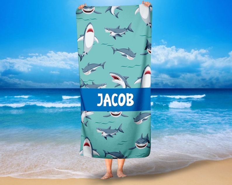 May include: A turquoise beach towel with a repeating pattern of cartoon sharks. The towel has a blue stripe across the middle with the name "JACOB" printed in white.