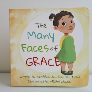 May include: The cover of a children's book titled "The Many Faces of GRACE". The cover features an illustration of a young girl with brown skin and brown hair wearing a green and yellow dress. The text is written in a colorful font.