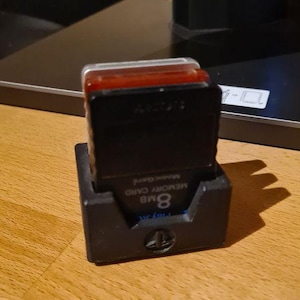 May include: A black plastic memory card holder containing an 8MB memory card with a black casing and a small red translucent top section. The memory card features the text 8MB MEMORY CARD printed in white on its front surface.