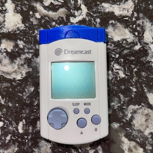 Peut inclure: Une console de jeu portable Sega Dreamcast blanche et bleue avec un bouton bleu sur le dessus et un écran affichant le logo Dreamcast. La console possède un pavé directionnel, quatre boutons et un bouton de veille et de mode.