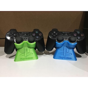 Op de afbeelding: Twee zwarte Sony PlayStation-gamecontrollers, elk rustend op een plastic standaard. De ene standaard is fel limoengroen en de andere is felblauw. Beide standaards zijn voorzien van het in reliëf gemaakte PlayStation-logo en het woord PlayStation.