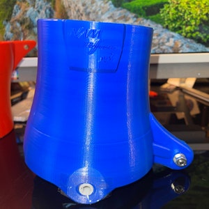 May include: A blue 3D printed part with the text "NAM Performance" on the side. The part has a hole in the bottom and a small arm extending out to the side.