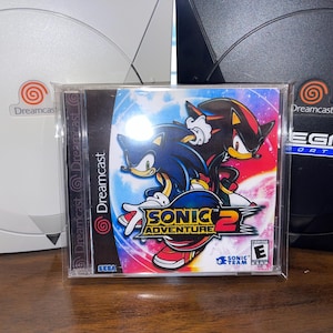 May include: A Sonic Adventure 2 video game case featuring Sonic and Shadow. The case is in front of a white and black Dreamcast console. The game case has a blue, red, and yellow color scheme.