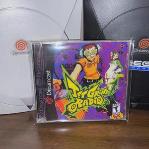 May include: A Jet Grind Radio video game case with a purple and green design, featuring cartoon characters. The case is in front of a white and a black Dreamcast console.
