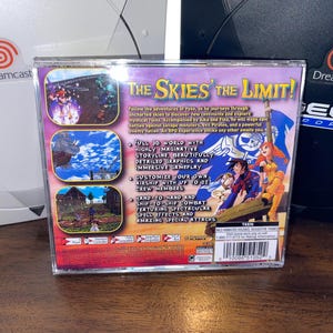 May include: Dreamcast game case for "The Skies of the Limit!" featuring colorful artwork of characters and scenes from the game. The case has text describing gameplay features and is set against a white and black Dreamcast console.
