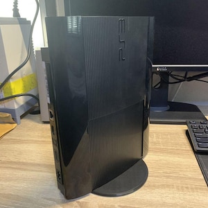 May include: A black PlayStation 3 Super Slim video game console standing vertically on a circular base. The console features a ribbed texture on the top section, a smooth glossy side panel, and the PS3 logo embossed on the upper right corner.