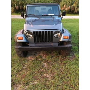May include: A light blue Jeep Wrangler with a black front bumper, a seven-slot vertical grille, round headlights, and amber turn signal lights. The vehicle features a black soft top, side mirrors, and two small auxiliary lights mounted near the windscreen.