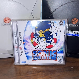 May include: A Sonic Adventure video game case with the Sega Dreamcast logo. The case features a colorful image of Sonic the Hedgehog. Two Dreamcast consoles are in the background, one white and one black.