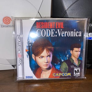 Peut inclure: Boîtier de jeu Sega Dreamcast pour Resident Evil Code: Veronica. La couverture présente deux personnages sur un fond bleu foncé et gris. Le boîtier du jeu est dans une pochette en plastique transparent. Le logo Sega Dreamcast est visible.