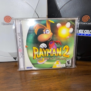 May include: A Rayman 2: The Great Escape video game case for the Sega Dreamcast console. The case features a colorful illustration of Rayman and a glowing orb. Two Dreamcast consoles, one white and one black, are in the background.