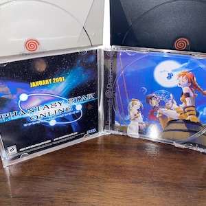 May include: A clear plastic case containing a Phantasy Star Online video game disc. The case is open, revealing the disc art with characters and a moonlit scene. The text "JANUARY 2001" is visible. Two Dreamcast consoles are in the background.