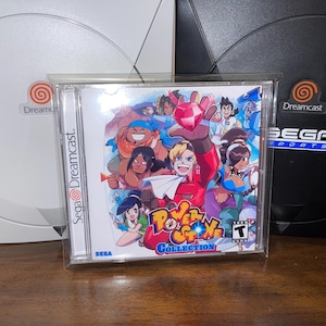 Könnte beinhalten: Eine Power Stone Collection Videospielhülle mit farbenfrohen Charakteren im Anime-Stil. Die Hülle befindet sich vor zwei Dreamcast-Konsolen, eine weiße und eine schwarze. Die Spielhülle hat das Sega-Logo.