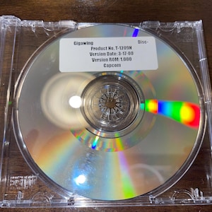 以下が含まれることがあります： 透明なプラスチックケースに入ったゲーム「ギガウイング」のCD-ROMディスク。ディスクは虹色の光屈折を持つ反射面を持っています。ラベルには、カプコンの製品とバージョンの情報が含まれています。