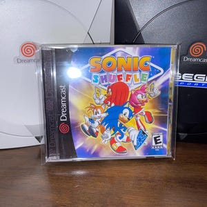 May include: A clear plastic case containing the Sonic Shuffle video game for the Sega Dreamcast. The game cover features Sonic the Hedgehog and other characters. Two Dreamcast consoles are in the background.
