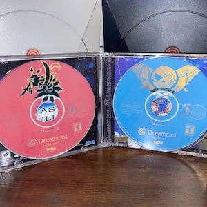 May include: Two Dreamcast game discs in clear cases. One disc is red with Japanese text and a stylized design. The other is blue with a gold emblem. Both discs have the Dreamcast logo.