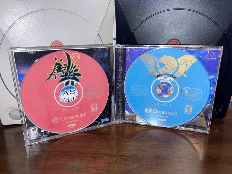 May include: Two Dreamcast game discs in clear cases. One disc is red with Japanese text and a stylized design. The other is blue with a gold emblem. Both discs have the Dreamcast logo.