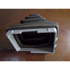 May include: A black rubber gear shift boot featuring a pleated, accordion-style design. The base of the boot is attached to a rectangular, light gray plastic frame with notched edges, designed for installation within a vehicle console.