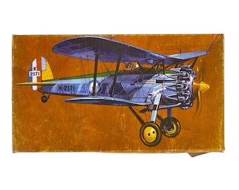 May include: A rectangular box featuring a vintage illustration of a blue biplane with a yellow propeller and wheels. The aircraft displays the text K-2171 on its side and tail, set against a solid, textured orange background with visible wear.