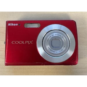 以下が含まれることがあります： 赤いニコンクールピクスデジタルカメラ。カメラは、前面に「NIKKOR 3X OPTICAL ZOOM」と「COOLPIX」の文字が入ったシルバーレンズを備えています。レンズの焦点距離は6.3〜18.9mm、絞りは1：3.1〜5.9です。