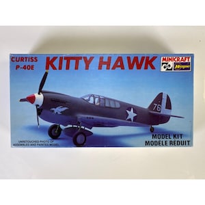 Può includere: Una scatola rettangolare per un kit di montaggio del Curtiss P-40E Kitty Hawk. La scatola presenta uno sfondo blu con l'illustrazione di un aereo grigio scuro, un ogiva dell'elica rossa e bianca, una stella bianca, il numero 76 e i loghi Minicraft Hasegawa.