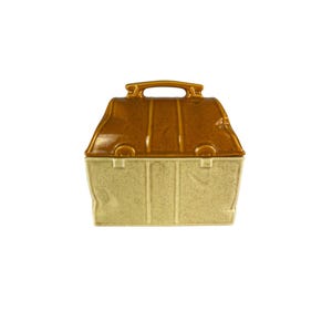 Pode incluir: Um recipiente de cerâmica de duas cores com uma alça, semelhante a uma lancheira vintage. A parte superior é marrom brilhante, enquanto a base é bege salpicado. O recipiente tem uma forma retangular com linhas verticais e uma alça na parte superior.