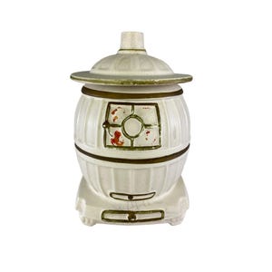 May include: A white ceramic jar with a lid, featuring a barrel-like shape with bronze-colored bands. The jar has a small window with red and green accents. The lid is light green with a textured pattern. The jar is a decorative item.