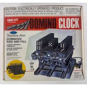 Puede incluir: Reloj Dominó vintage en su caja original. El reloj presenta un diseño único donde las fichas de dominó suben y bajan para indicar la hora. La caja tiene el texto "Snap-Fit Electric Domino Clock" y "Fascinating Action Dominoes Rise and Fall".