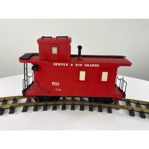 May include: A red toy caboose with the text "DENVER & RIO GRANDE" and the number "701" printed on the side. The caboose has a black roof and is sitting on a section of toy train tracks.