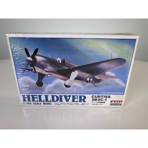 May include: A rectangular box for a 1/144 scale model kit of a Curtiss SB2C-1 Helldiver aircraft. The box features a blue sky background with white clouds and a detailed illustration of the dark gray plane in flight with its landing gear down.