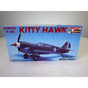 Puede incluir: Una caja de cartón rectangular para un kit de modelo a escala Curtiss P-40E Kitty Hawk. La caja presenta un fondo azul con una ilustración de un avión de combate gris oscuro con una estrella blanca, una punta de hélice roja y el número 76.
