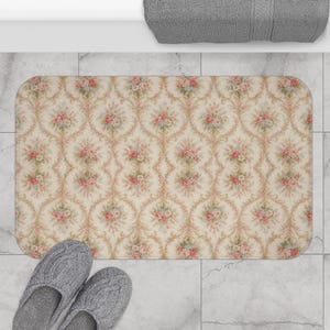 May include: A rectangular bath mat with a floral pattern in shades of pink, green, and beige. The mat features a repeating design of roses and decorative scrollwork. Gray slippers are at the bottom of the mat.