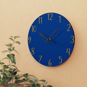 May include: A blue round clock with gold numbers and black hands. The clock is on a beige wall.