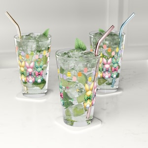 May include: Three clear glass tumblers with colorful Easter bunny and egg designs. Each glass has a straw and is filled with ice and a clear liquid. The glasses are on a white surface.