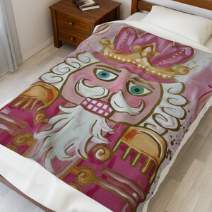 May include: A pink and gold nutcracker blanket with a whimsical design. The blanket features a detailed illustration of a nutcracker with a crown, mustache, and a stern expression. The color palette includes shades of pink, gold, and white.