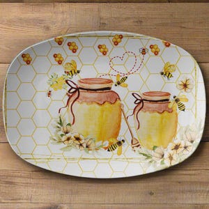 May include: Oval serving platter with a bee and honey theme. The white surface features a honeycomb pattern and illustrations of honey pots, bees, and flowers. The rim has a gold border. The honey pots have brown lids and are tied with brown ribbons.