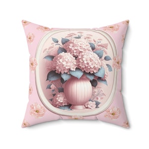 May include: A pink throw pillow with a floral design. The pillow features a bouquet of pink hydrangeas in a pink vase, surrounded by a white oval frame. The background is a light pink with a repeating pattern of smaller pink flowers.